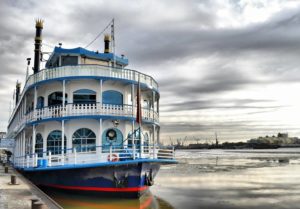 a paddle steamer beat sits docked at a river dock. The sky is cloudy but bright since the clouds are thin. the steam boat is a two smokestack, three story blue white and red vessel.